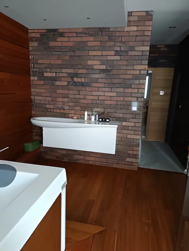 Koupelna se stěnou v imitaci červených cihel za bílým umyvadlem a dřevěnou podlahou